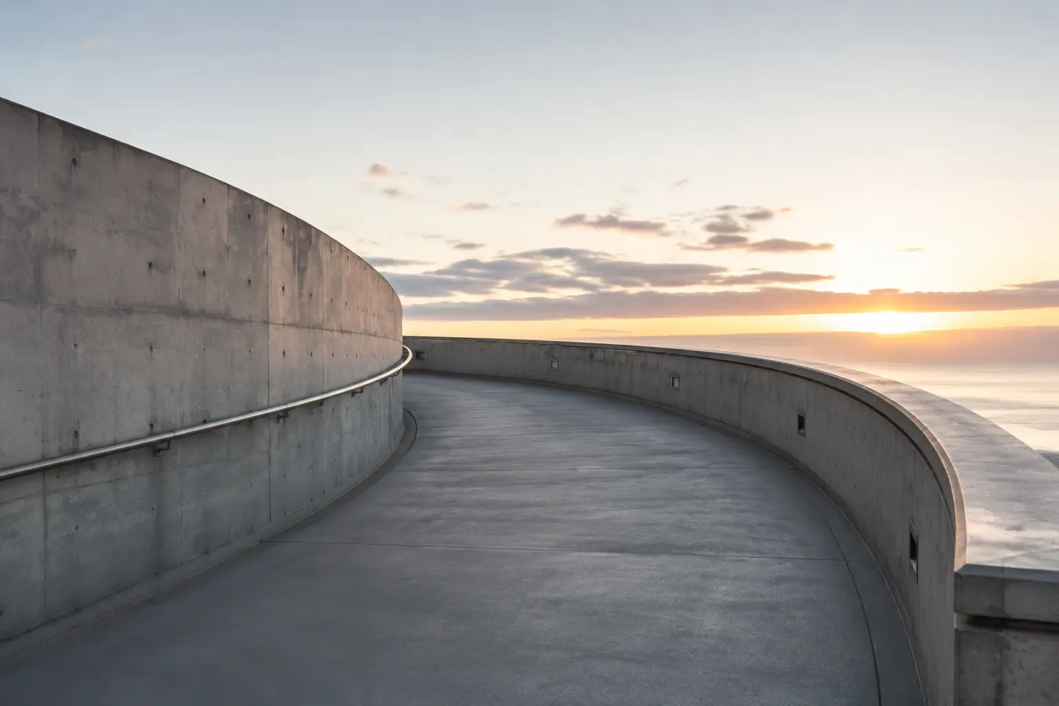 Empty curved concrete passage opening toward a warm dawn horizon